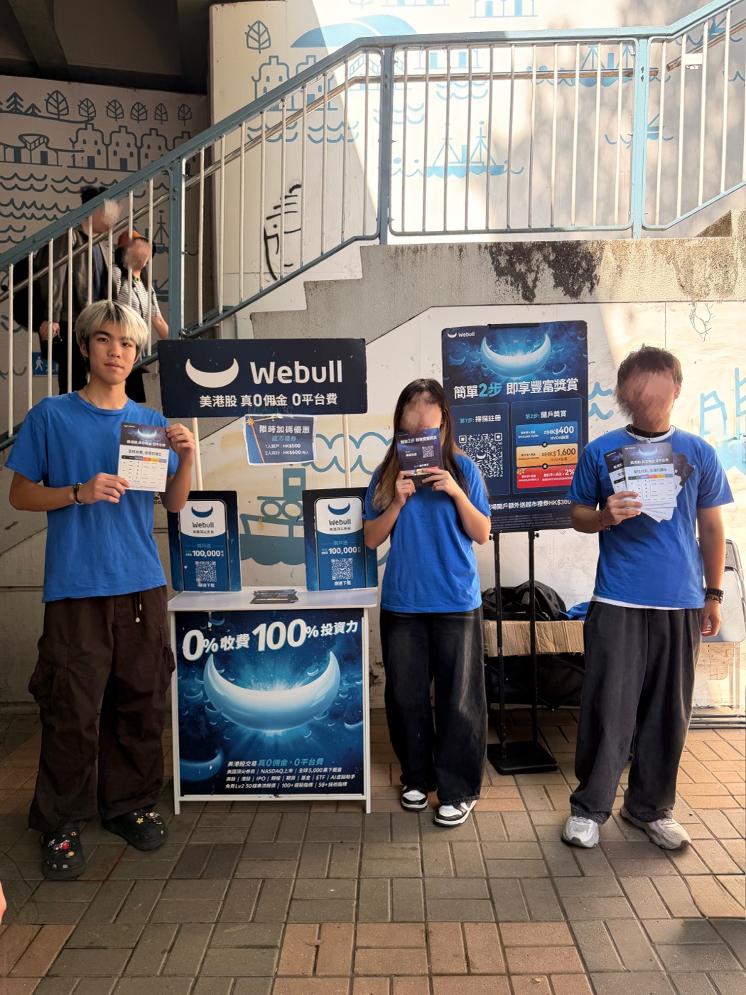 Three promoters in blue Webull T-shirts holding flyers beside a Webull booth with QR-code sign-up boards, under a stairway.