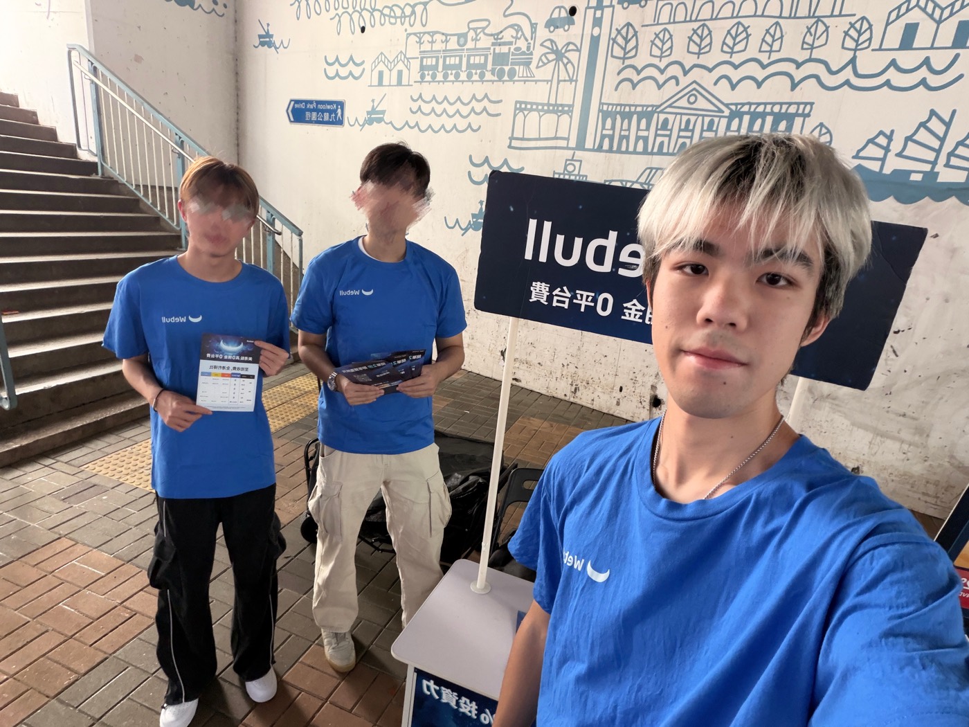 Wyatt Lo taking a selfie with two teammates in Webull T-shirts at a roadshow by the Kowloon Park Drive underpass.